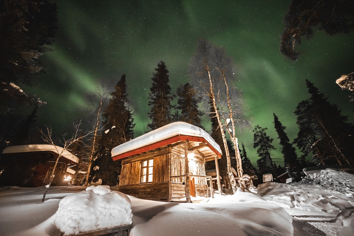 Lumihuntuinen puumökki, jonka katto on peittynyt lumeen. Mökkiä ympäröi lumi ja puut, ja sen yllä loistaa tähtitaivas sekä revontulet.