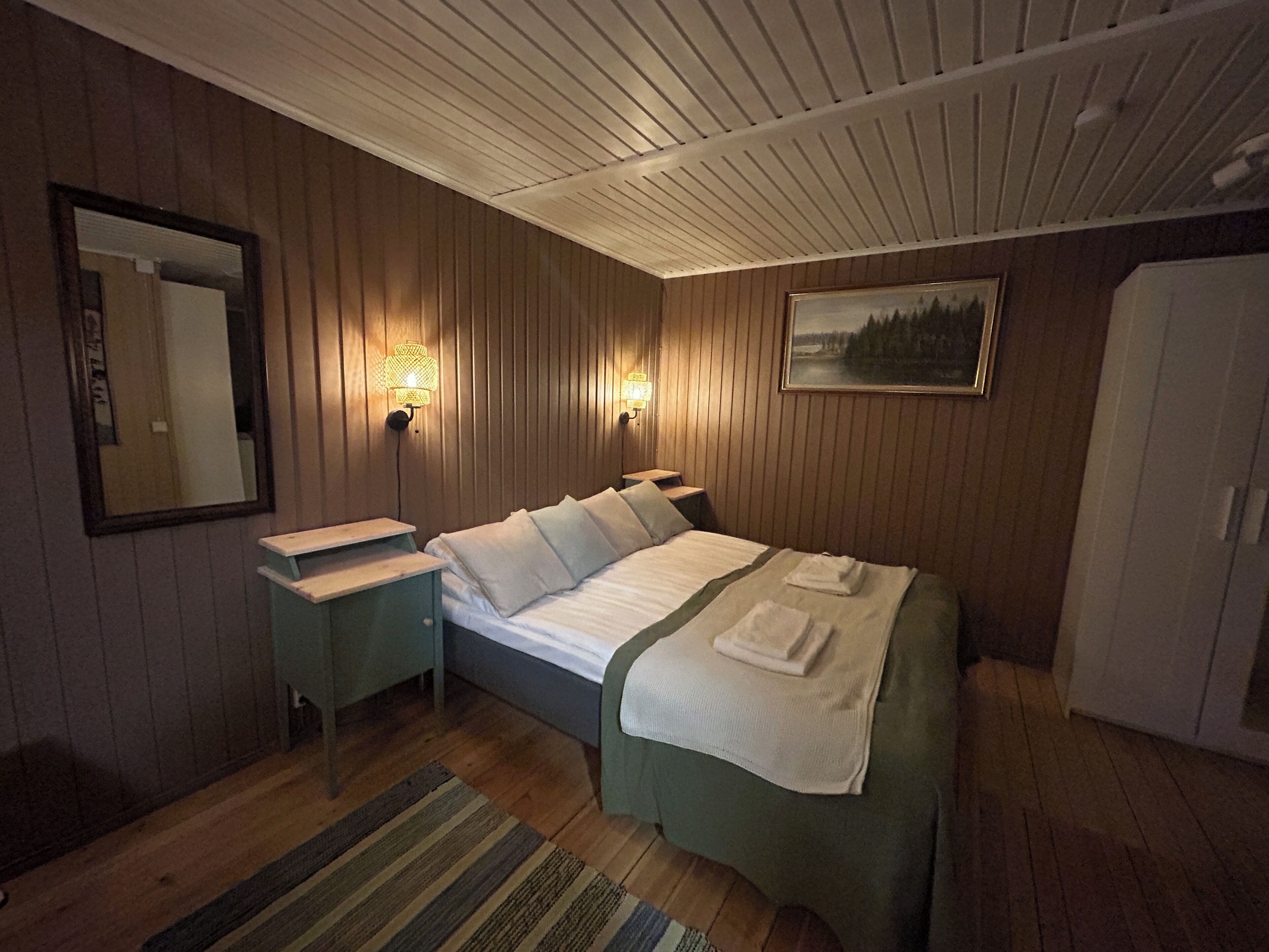 Een hotelkamer met een bed, nachtkastjes, een spiegel, wandlampen, een schilderij en een kledingkast.