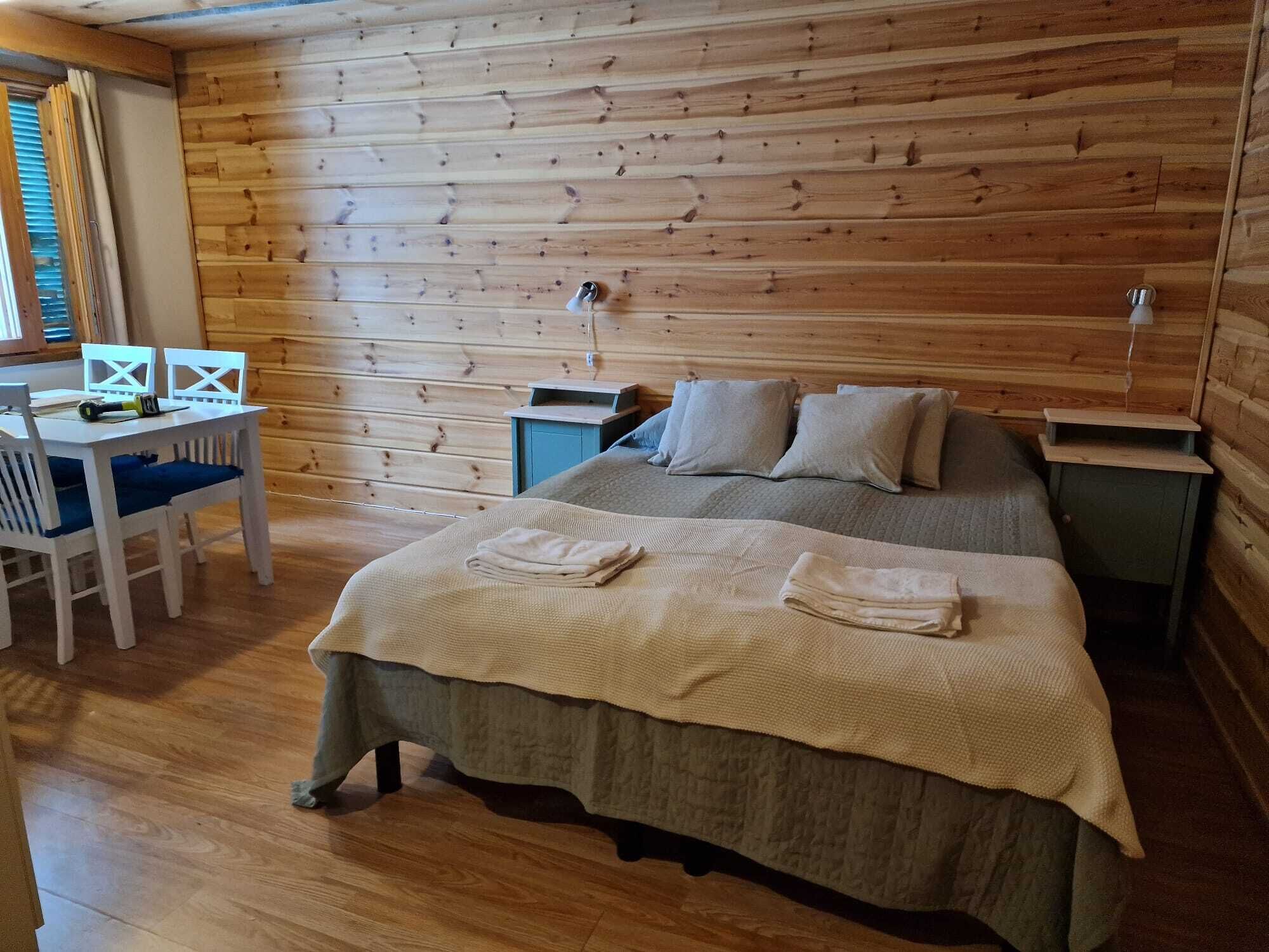 A cozy room with a wooden log wall, a bed with a beige blanket, a desk with a chair, and a nightstand.