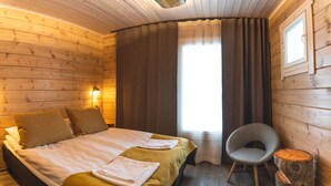 A wooden room with a bed, two pillows, a chair, a nightstand, and a window with curtains. - Tankavaara Gold Village (Sodankyla)