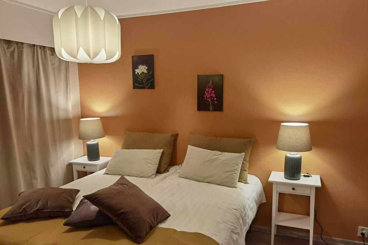 Hotellihuoneessa on sänky, kaksi yöpöytää, molemmilla pöydillä on lamppu ja seinällä on taulu.