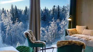 Una habitación con un ventanal grande con vistas a un bosque, sillas tapizadas de verde, una mesa con individuales tejidos y una lámpara en una mesita auxiliar.