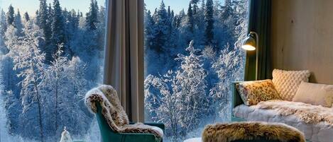 Quarto com uma grande janela com vista para uma floresta, cadeiras estofadas em verde, mesa com jogos americanos trançados e abajur sobre uma mesinha lateral.