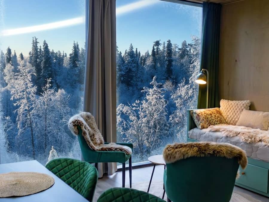 Ein Zimmer mit großem Fenster und Blick auf einen Wald, mit grünen Polstersesseln, einem Tisch mit gewobenen Platzsets und einer Lampe auf einem Beistelltisch.
