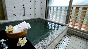 Un espace détente avec une baignoire en bois, un récipient en bois contenant des serviettes et une fenêtre donnant sur un paysage agréable.