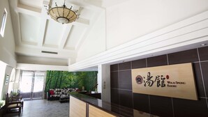 Un espace d’accueil avec un comptoir de réception en bois, une enseigne murale indiquant « Wulai Spring Resort », un plafond orné d’un lustre et une vue sur un mur végétal, visible à travers une porte vitrée.