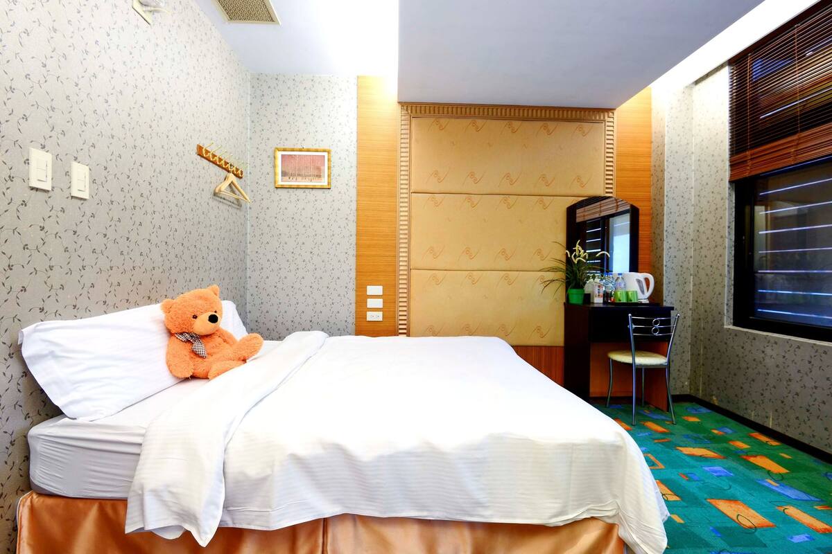 Een hotelkamer met een bed, een teddybeer, een bureau met stoel, een raam met jaloezieën en een behang met patroon.