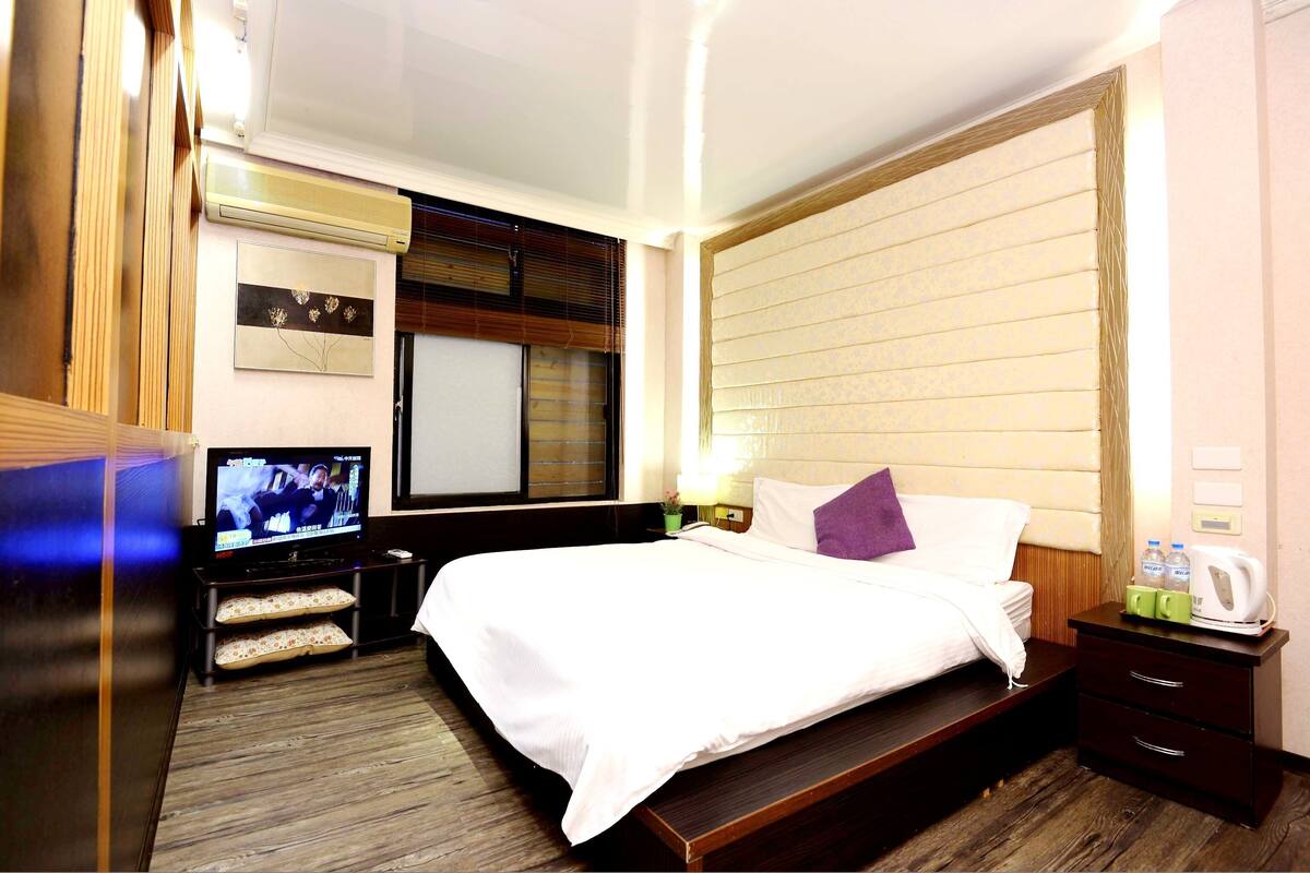 Een hotelkamer met een bed, een televisie, een nachtkastje met lamp en een raam met jaloezieën.
