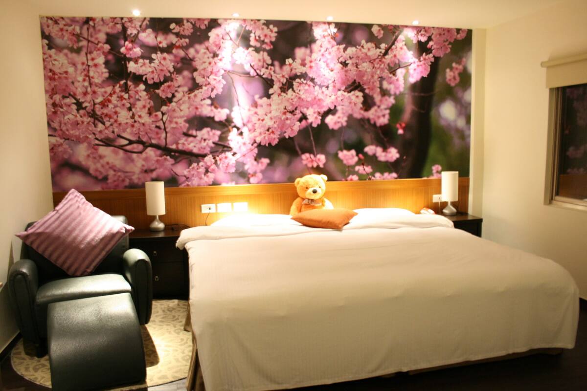 Een hotelkamer met een groot bed, een hoofdeinde met bloemenbehang, een nachtkastje met een lamp en een stoel met een kussen.