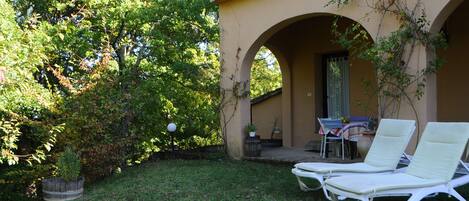 A patio with two white lounge chairs, a table with chairs, and a small garden area with a tree and a planter.