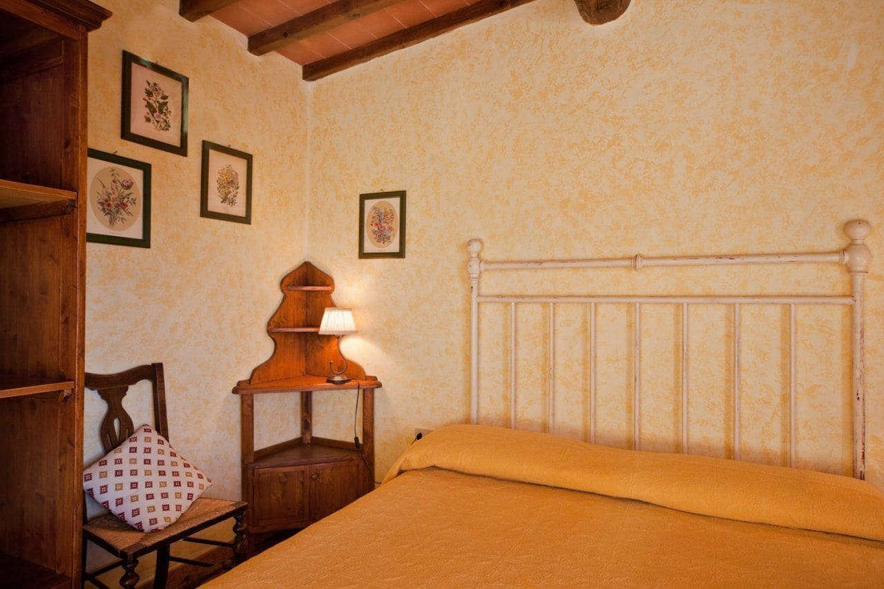 Une chambre à coucher avec une tête de lit en bois, un lit recouvert d’une couverture jaune, une table de chevet en bois avec une lampe, et une chaise agrémentée d’un coussin à motifs.