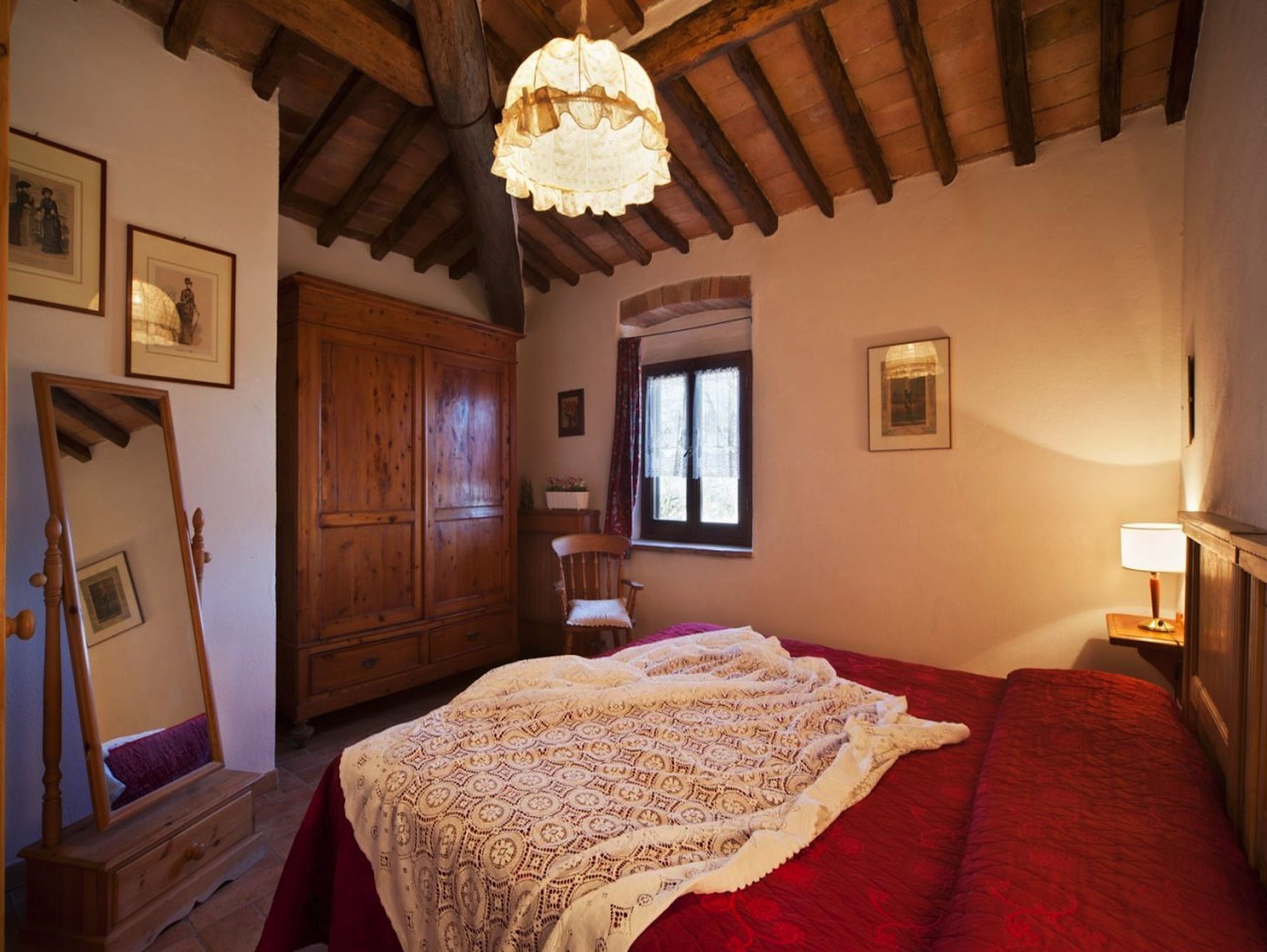 Quarto com um guarda-roupa de madeira grande, cama com manta vermelha, candelabro e quadros nas paredes.