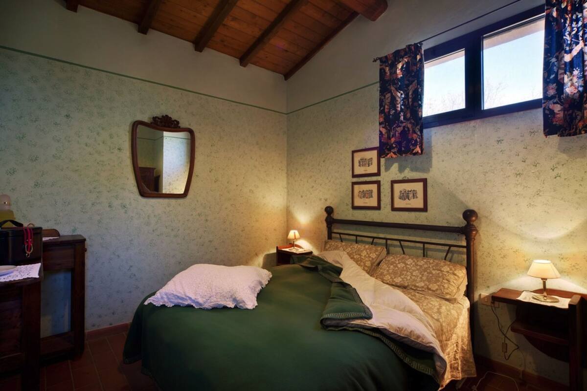 A bedroom with a wooden ceiling, floral wallpaper, a bed with green and patterned bedding, a nightstand with a lamp, and a window with floral curtains.