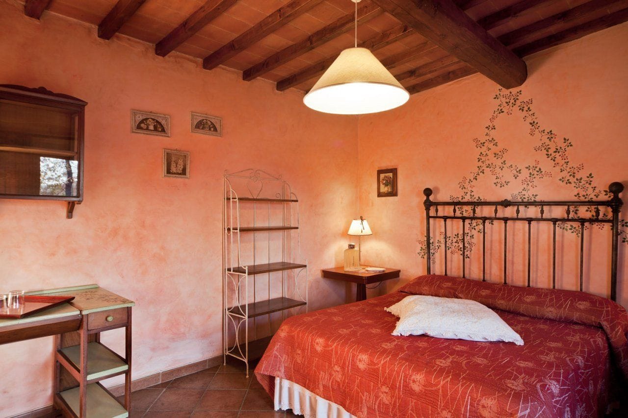A bedroom with a wooden ceiling, a bed with a floral bedspread, a bedside table with a lamp, and a metal shelf unit.