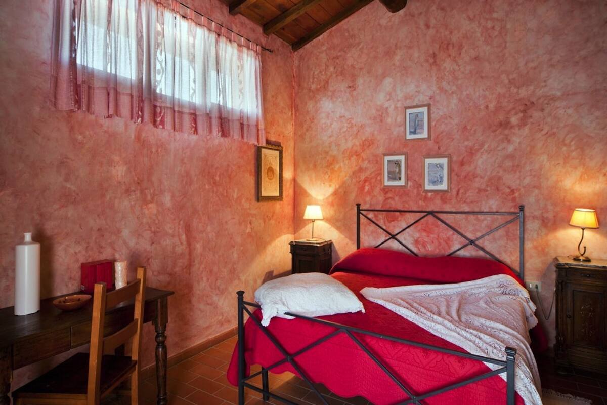 A bedroom with a wooden ceiling, a metal bed frame, a red bedspread, a wooden chair, a bedside table with a lamp, and a wall with three framed pictures.