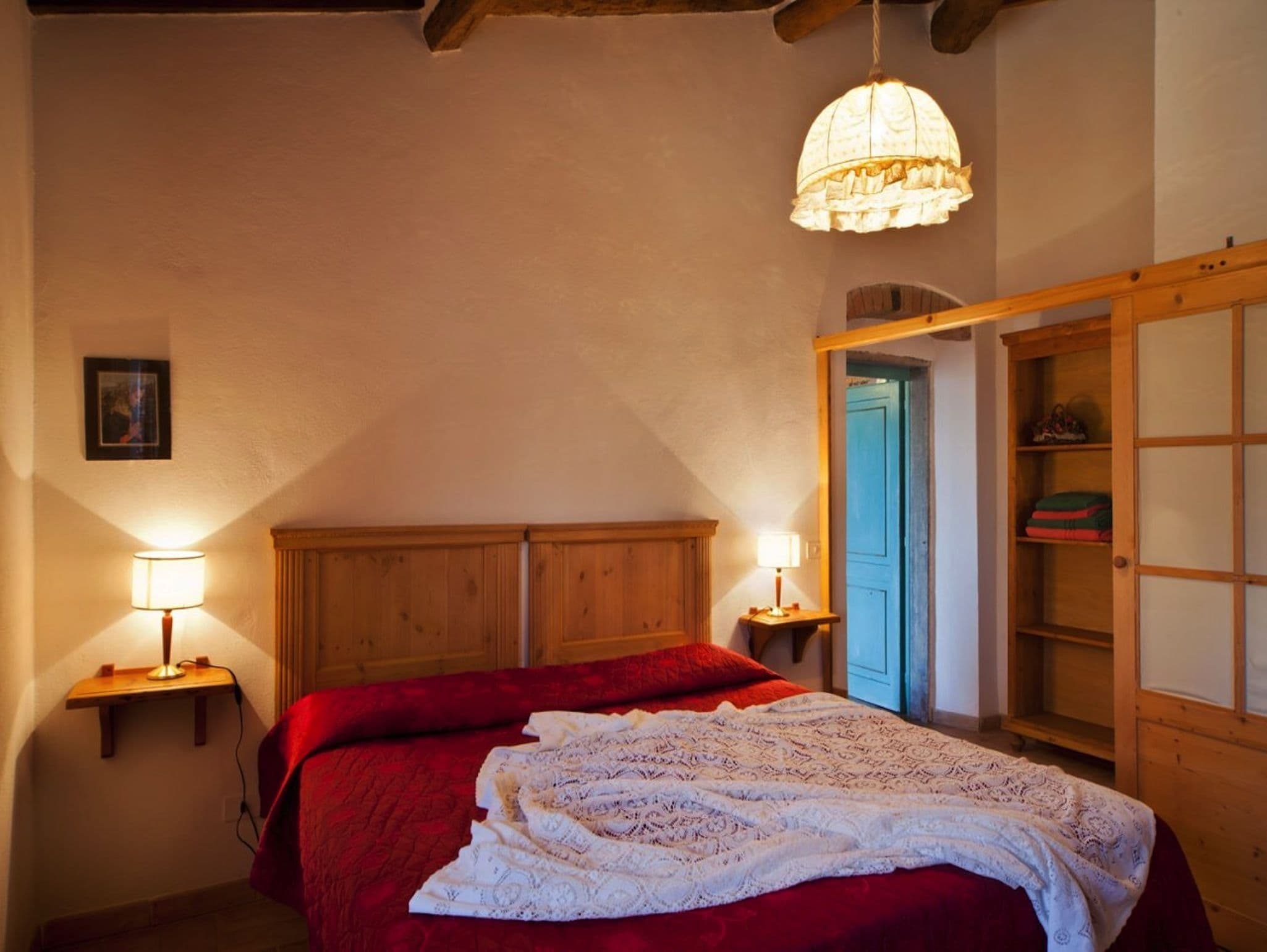 A bedroom with a wooden headboard, a red bedspread, a white lace throw, two bedside lamps, and a wooden wardrobe.