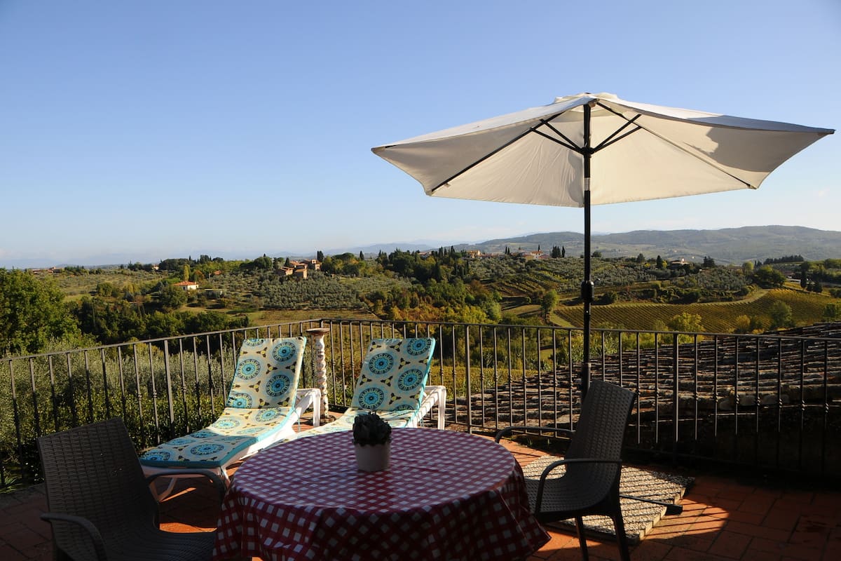A terrace with a table and chairs, a large umbrella, and a scenic view of rolling hills and vineyards.