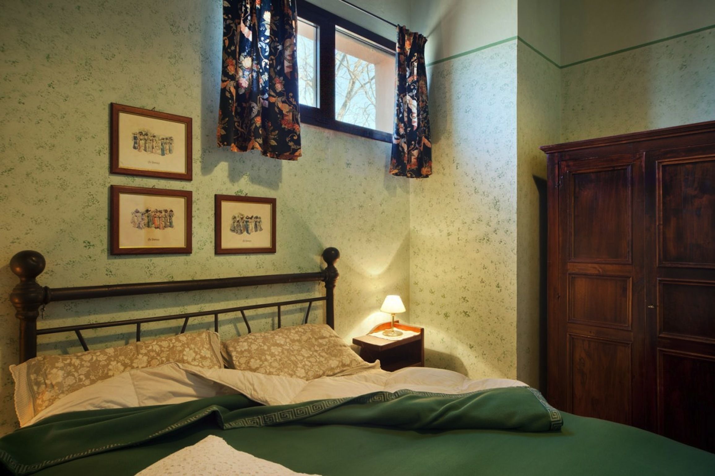A bedroom with a bed, a nightstand with a lamp, a wardrobe, and three framed pictures on the wall.