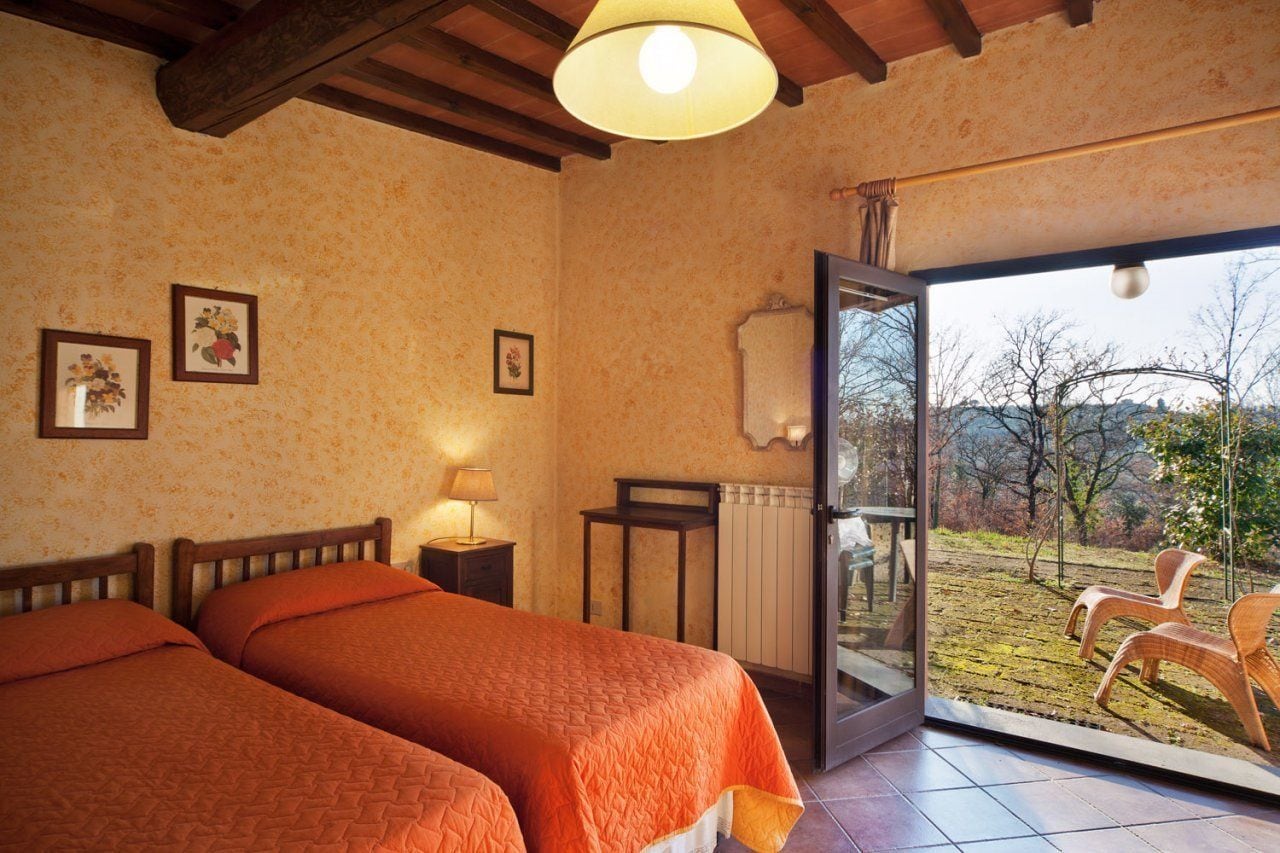 A room with two beds, a lamp, framed pictures, and a view of an outdoor area with chairs.
