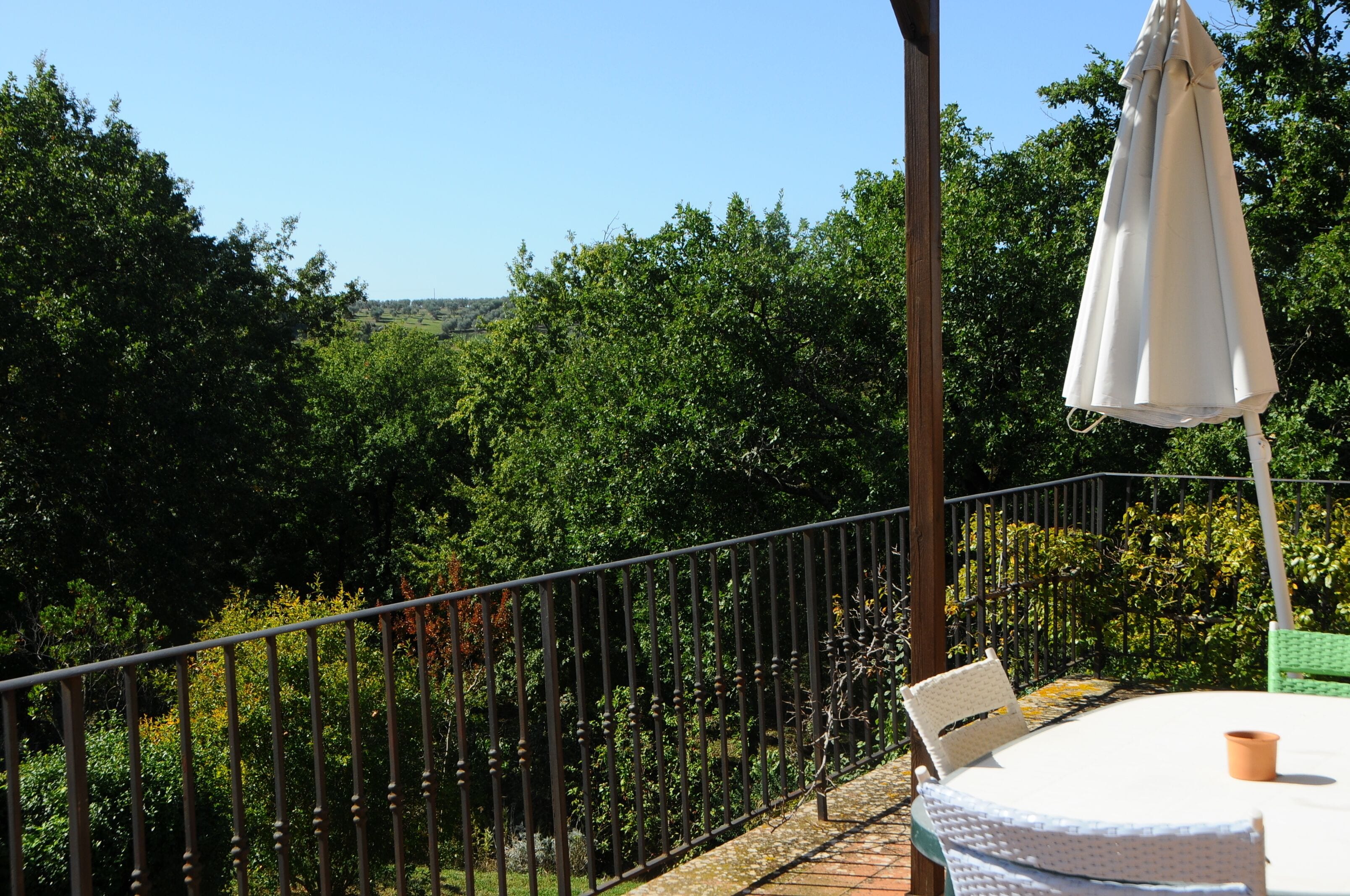 Een balkon met een tafel, stoelen, een witte parasol en een metalen hekwerk.