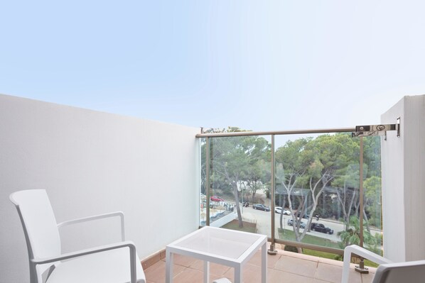 A balcony with a white chair, a small table, and a view of a street with trees and parked cars.
