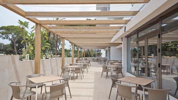 Un espacio al aire libre con varias mesas y sillas, una pérgola de madera en la parte superior y puertas de vidrio que dan a una zona de piscina.