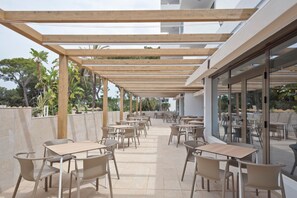 An outdoor patio area with multiple tables and chairs, wooden pergola overhead, and glass doors leading to a pool area. - Hotel Best Delta (Llucmajor)
