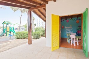 A children's play area with a slide and climbing structure, a wooden pergola, and a wall with colorful decorations and a cartoon character. - Hotel Best Delta (Llucmajor)