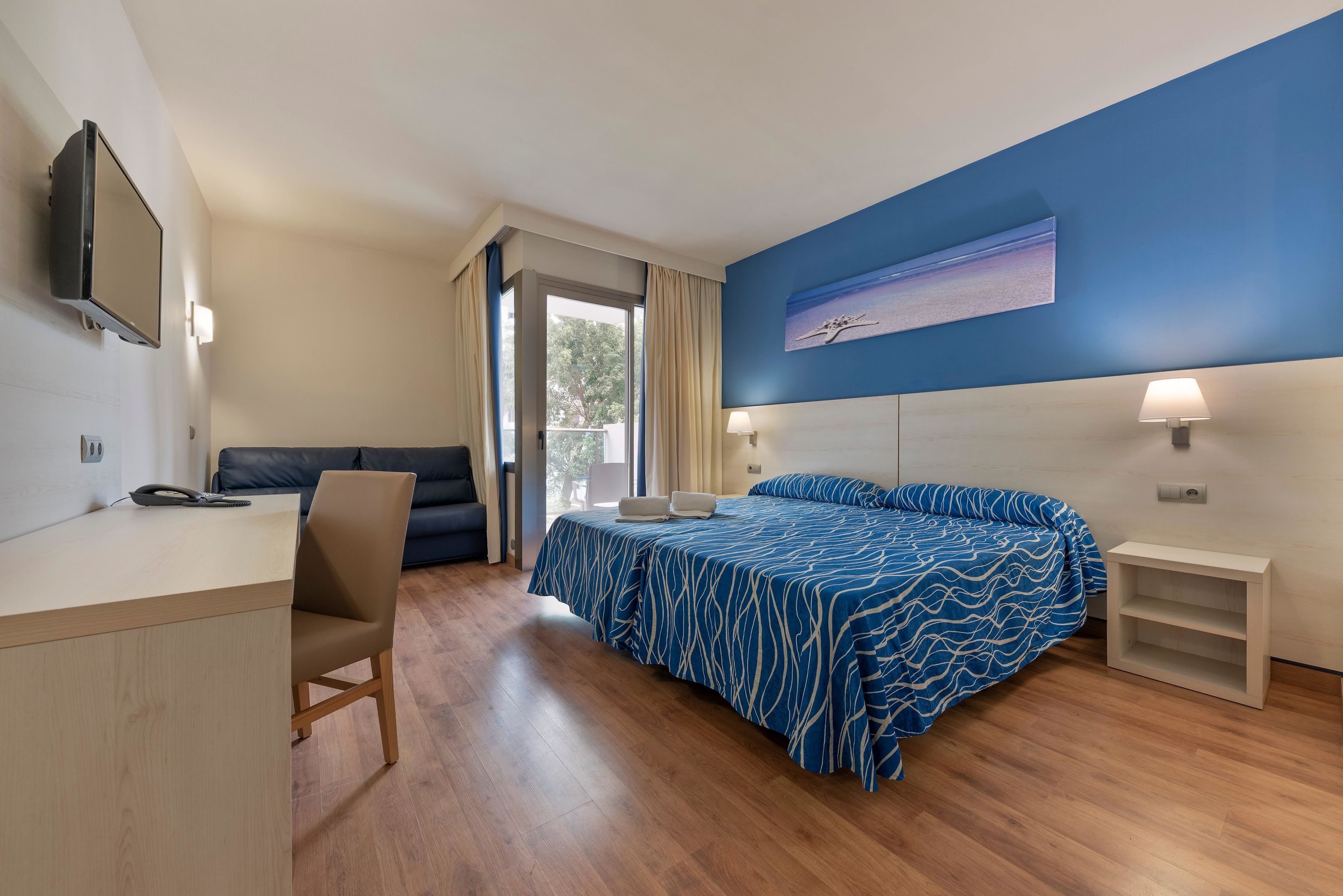 Habitación de hotel con cama, escritorio con silla, televisor montado en la pared y ventana con cortinas.