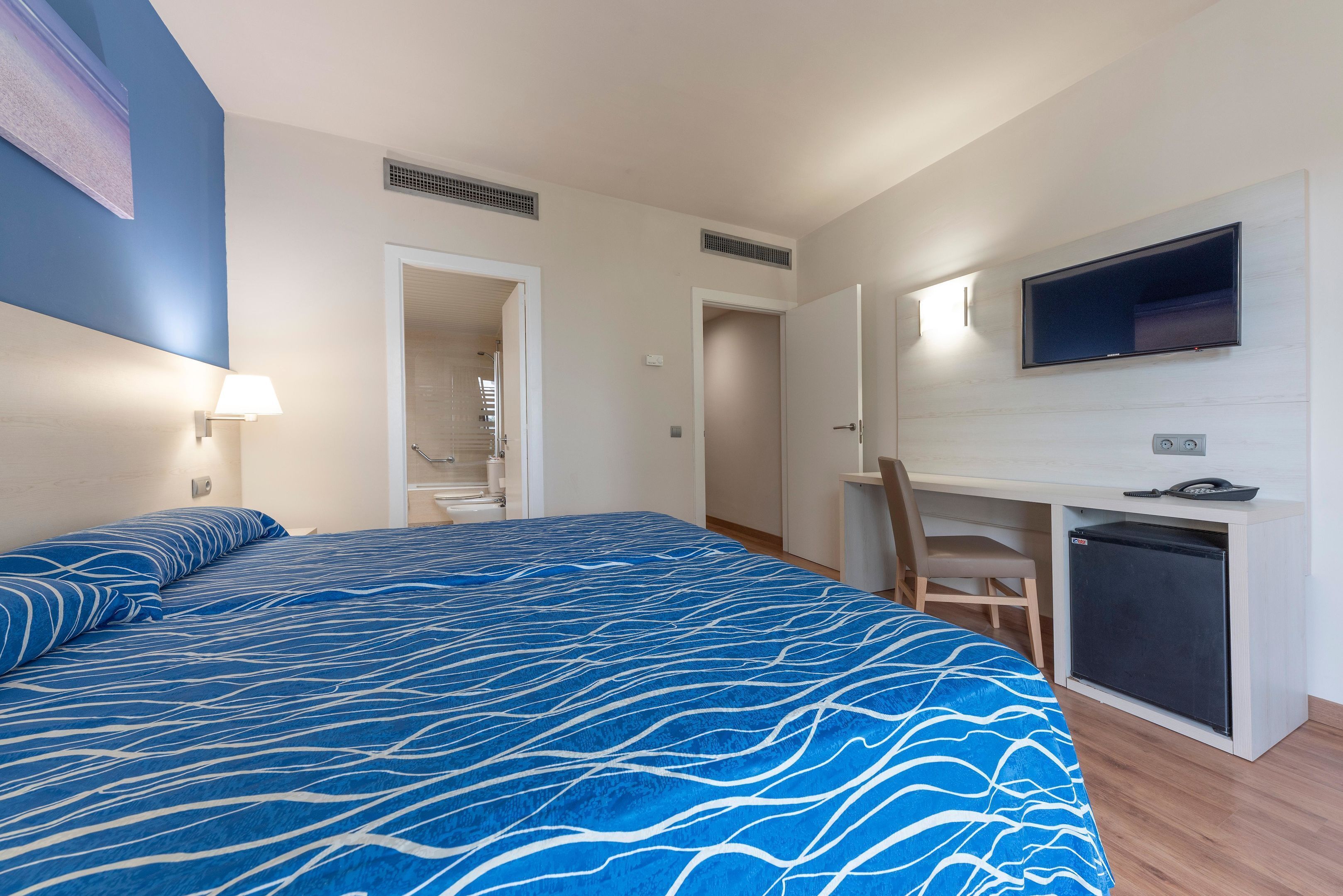 Habitación de hotel con cama, escritorio, televisor y refrigerador pequeño.
