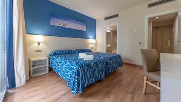 Una habitación de hotel con cama, mesitas de noche, una silla, un cuadro en la pared y una puerta que da a otra habitación.