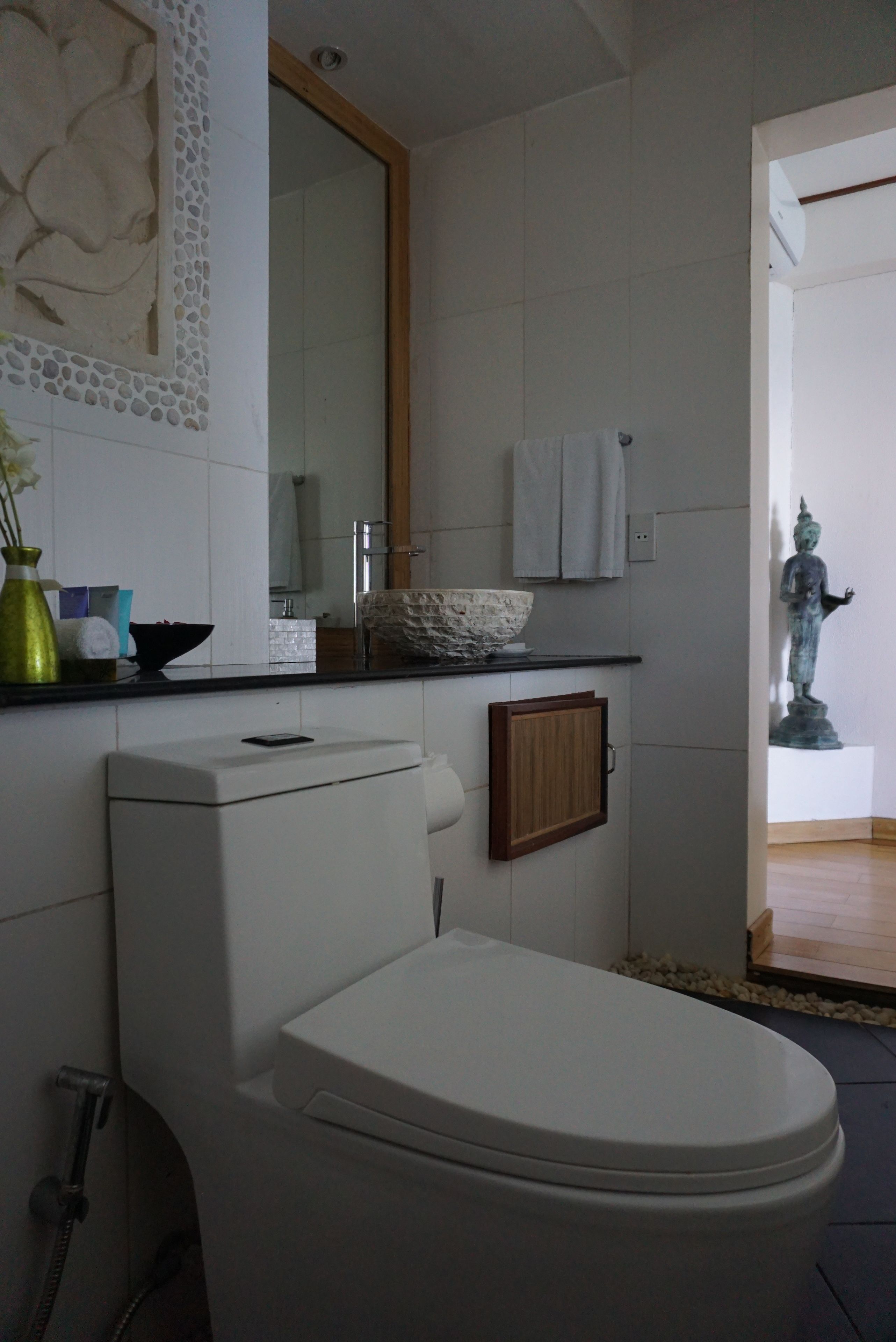 Et moderne badeværelse med et hvidt toilet, en vask med en dekorativ skål, et spejl og en statue i hjørnet.