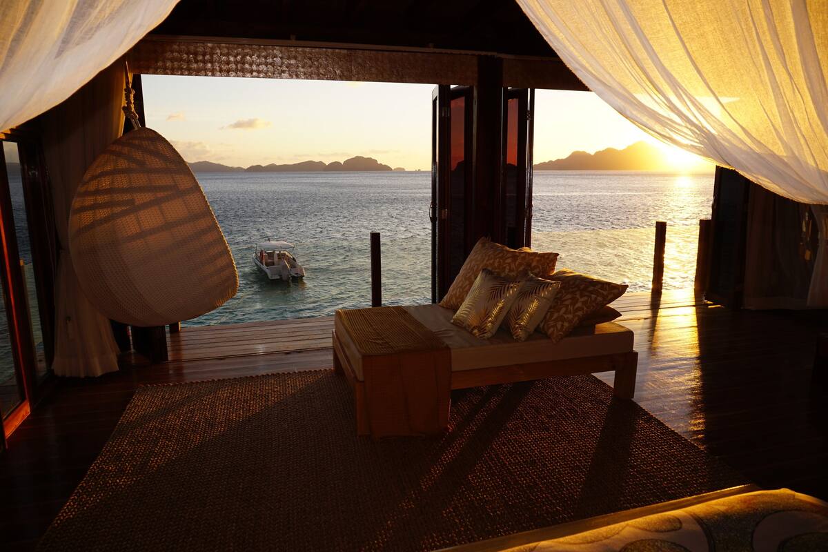 A room with a bed, a view of the sea, and a boat in the distance.
