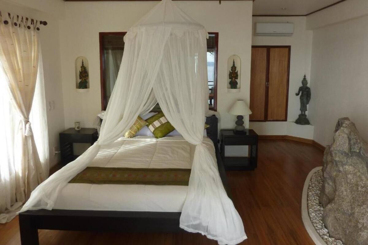 A bedroom with a canopy bed, wooden flooring, a ceiling fan, and statues on the walls.