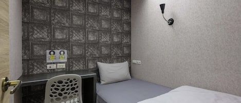 A small, compact room with a bed, a desk, and a chair. There is a wall-mounted light fixture and a power outlet on the wall.