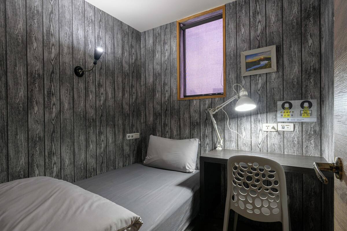 A room with a bed, a desk, a chair, a window, and a wall-mounted light fixture.