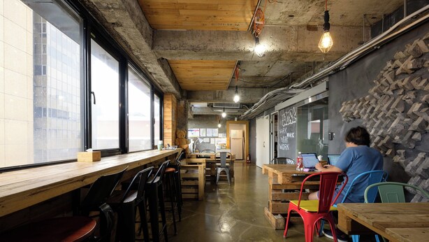 A café with wooden tables and chairs, a person working on a laptop, and a stone wall. - Work Inn TPE - Hostel (Taipei)
