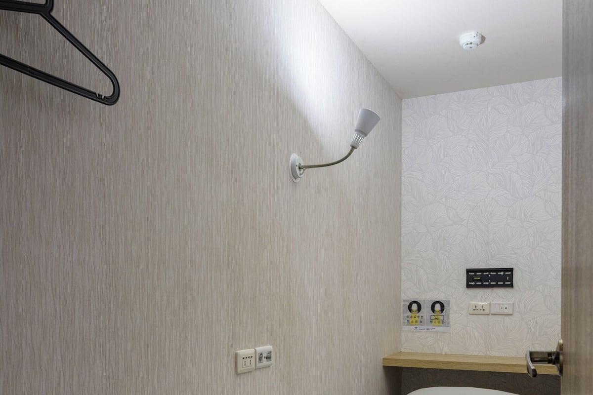 A small, single-bed room with a wooden bed frame, a bedside table with a lamp, a wall-mounted heater, and a small shelf with a mirror and a power outlet.