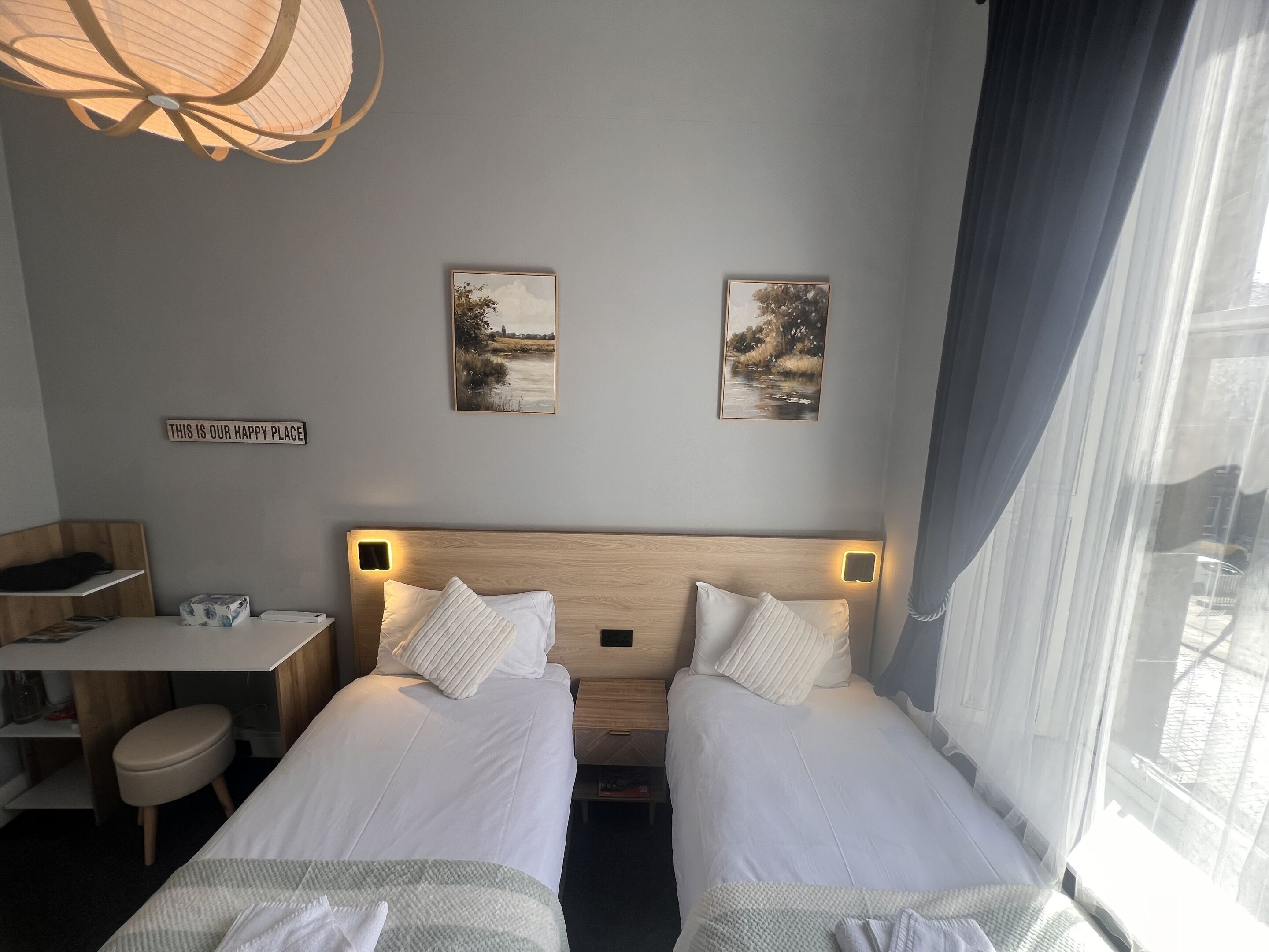 Une chambre d’hôtel avec deux lits, un bureau, une chaise et une fenêtre avec des rideaux. Il y a deux tableaux encadrés au mur et une pancarte sur laquelle on peut lire : « ICI, NOUS SOMMES HEUREUX ».