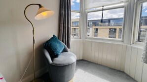 Una habitación de esquina con un sillón gris, una lámpara de pie y un amplio ventanal con vistas a edificios.