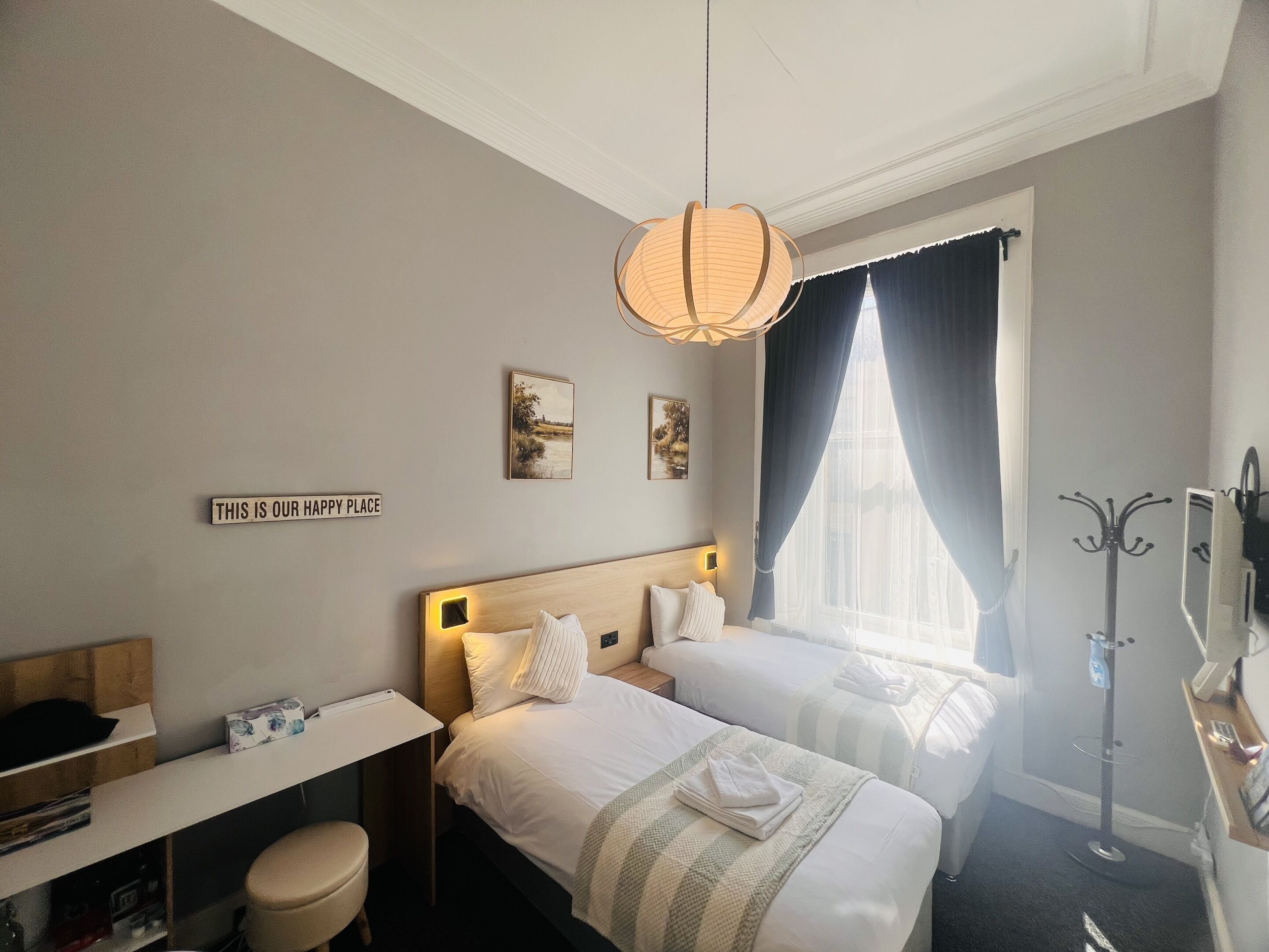 Habitación de hotel con dos camas, un escritorio, una silla, perchero y obras de arte en las paredes.