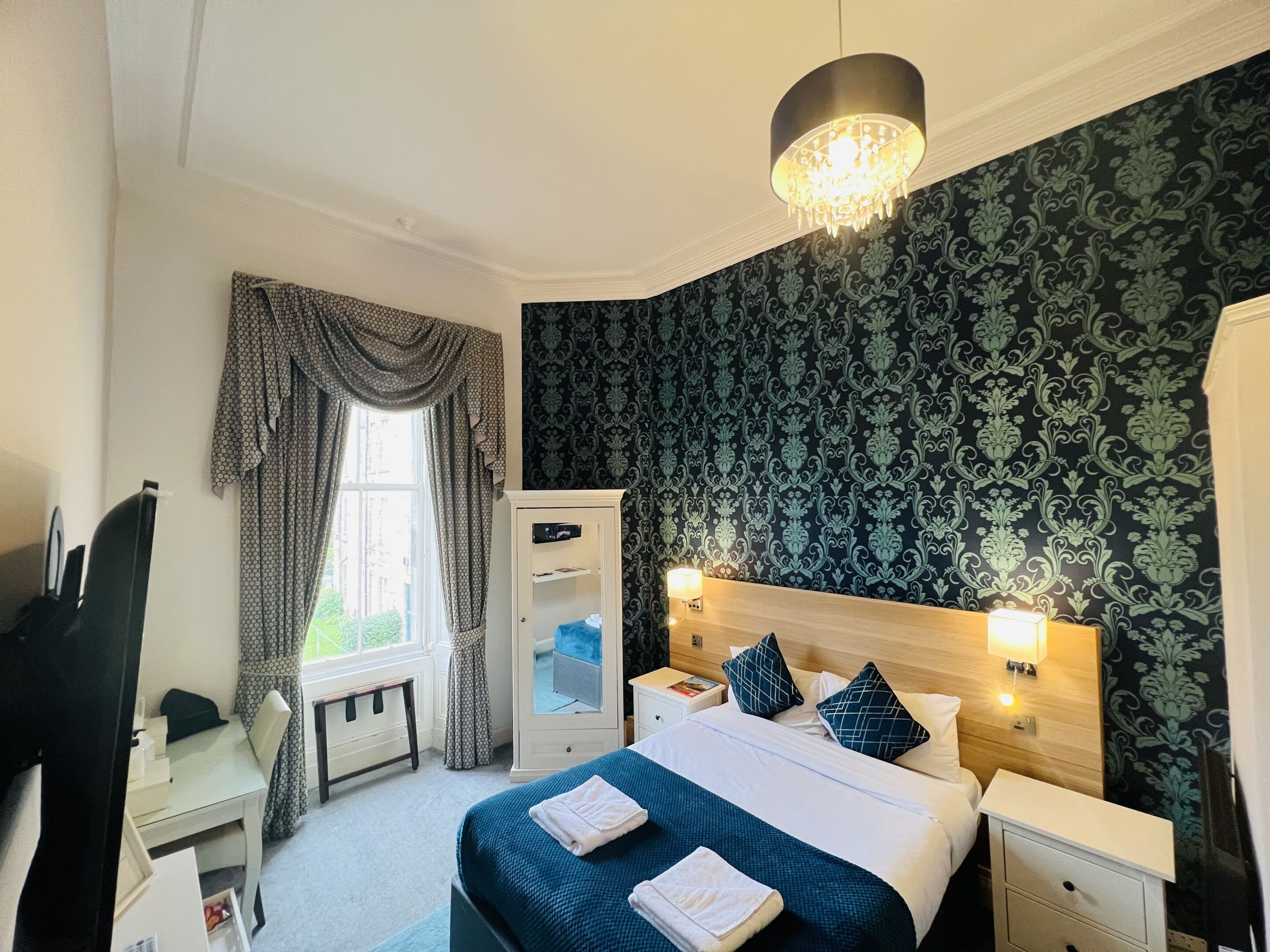 A hotel room with a bed, bedside tables, a desk, a chair, a wardrobe, and a window with curtains.