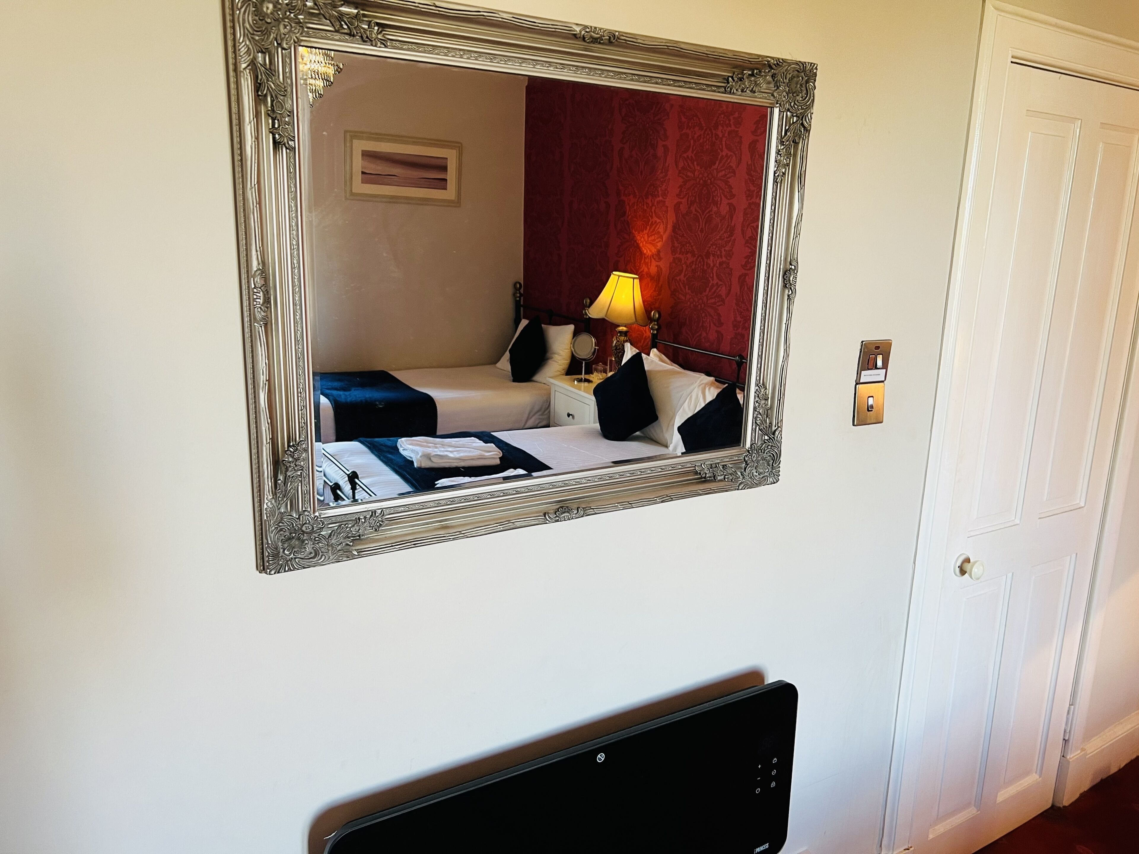 Hotellihuone, jossa on suuri, koristeellinen peili, mustavalkoraidallinen päiväpeite sängyssä, yöpöytä lampun kanssa ja valkoinen ovi.