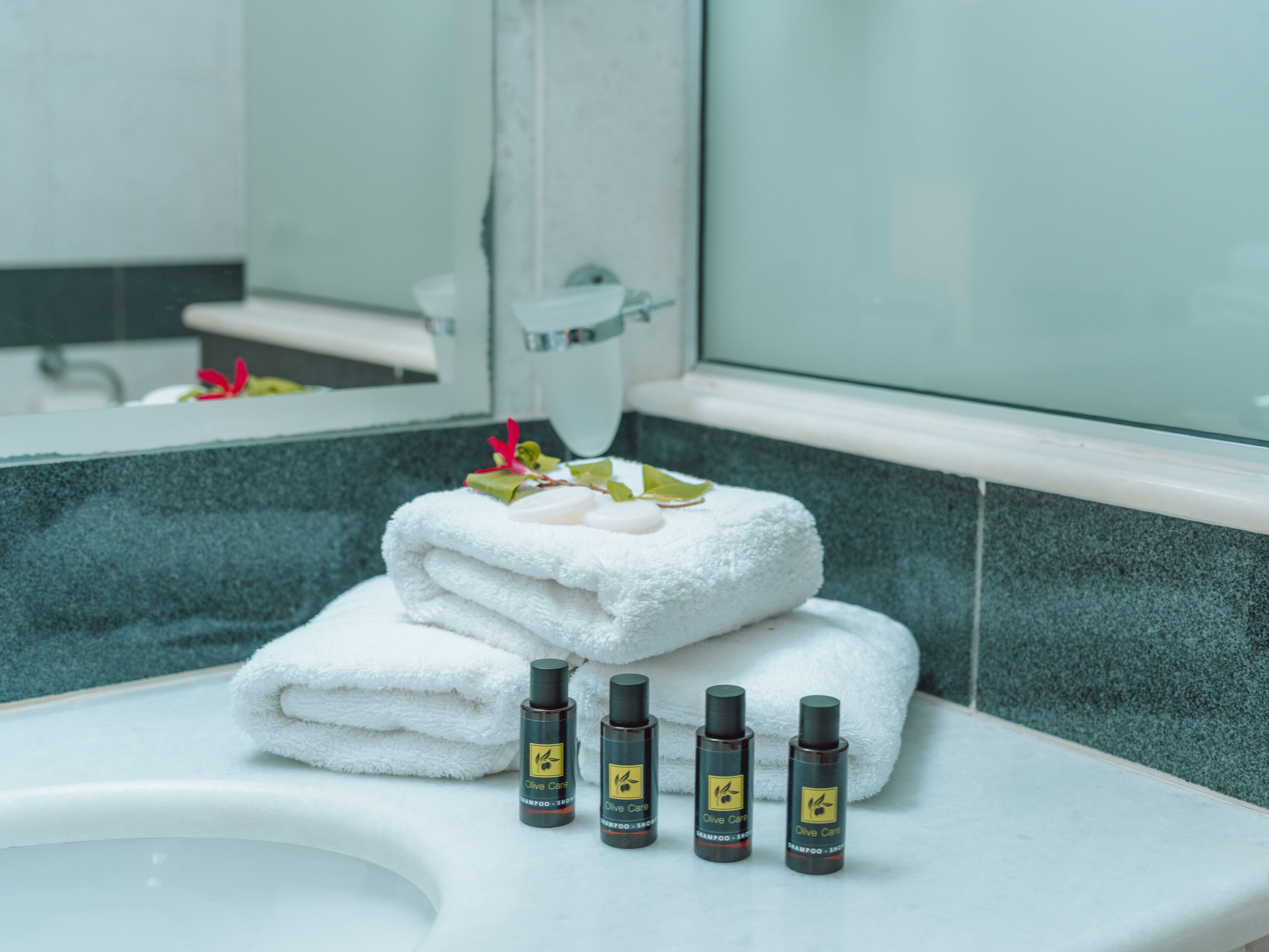 Un baño con un lavamanos blanco, una pila de toallas blancas y tres botellas de champú Olive Care.