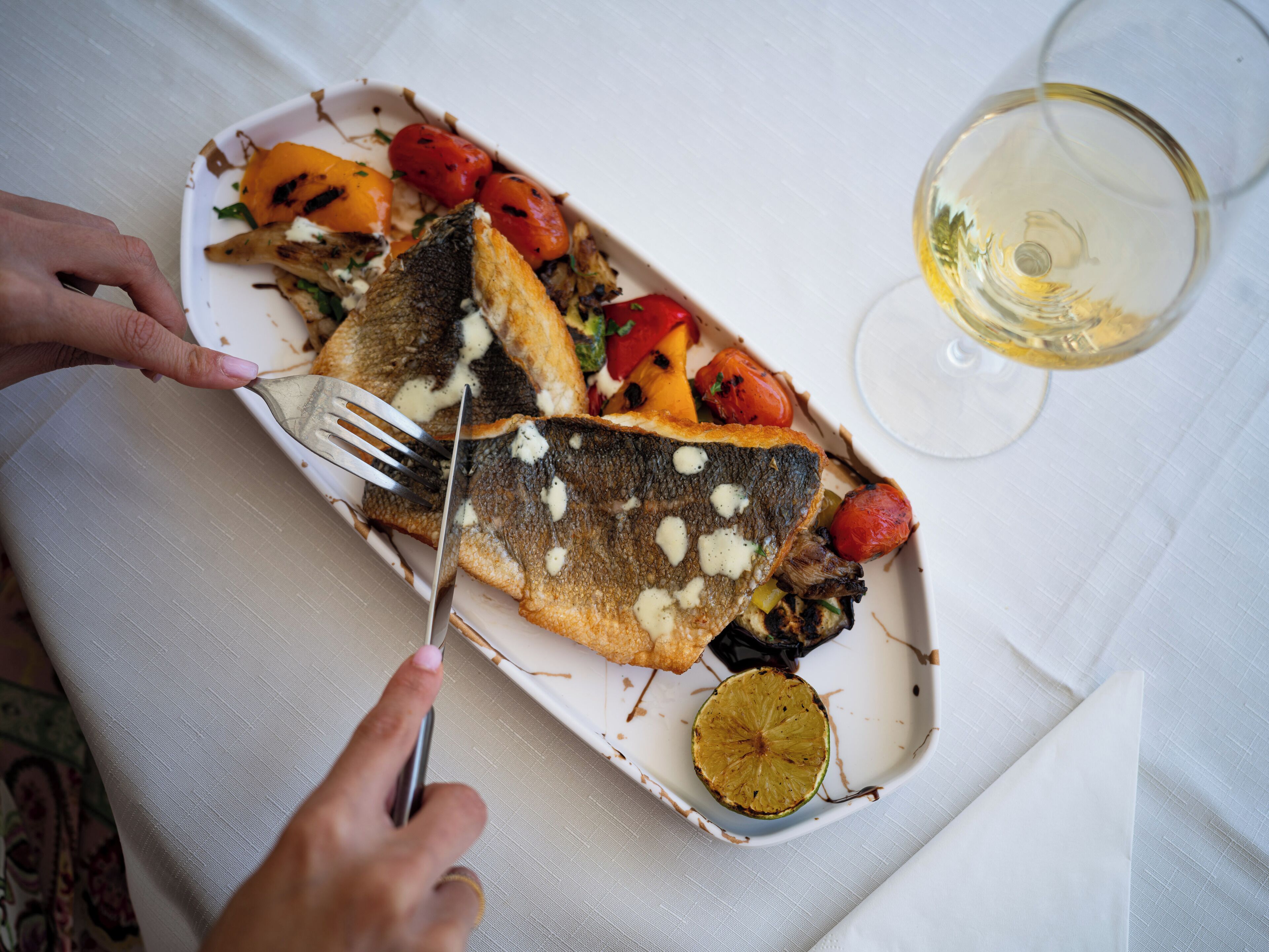 Un plat de menjar amb peix, verdures i una làmina de llimona, acompanyat d'un got de vi blanc.