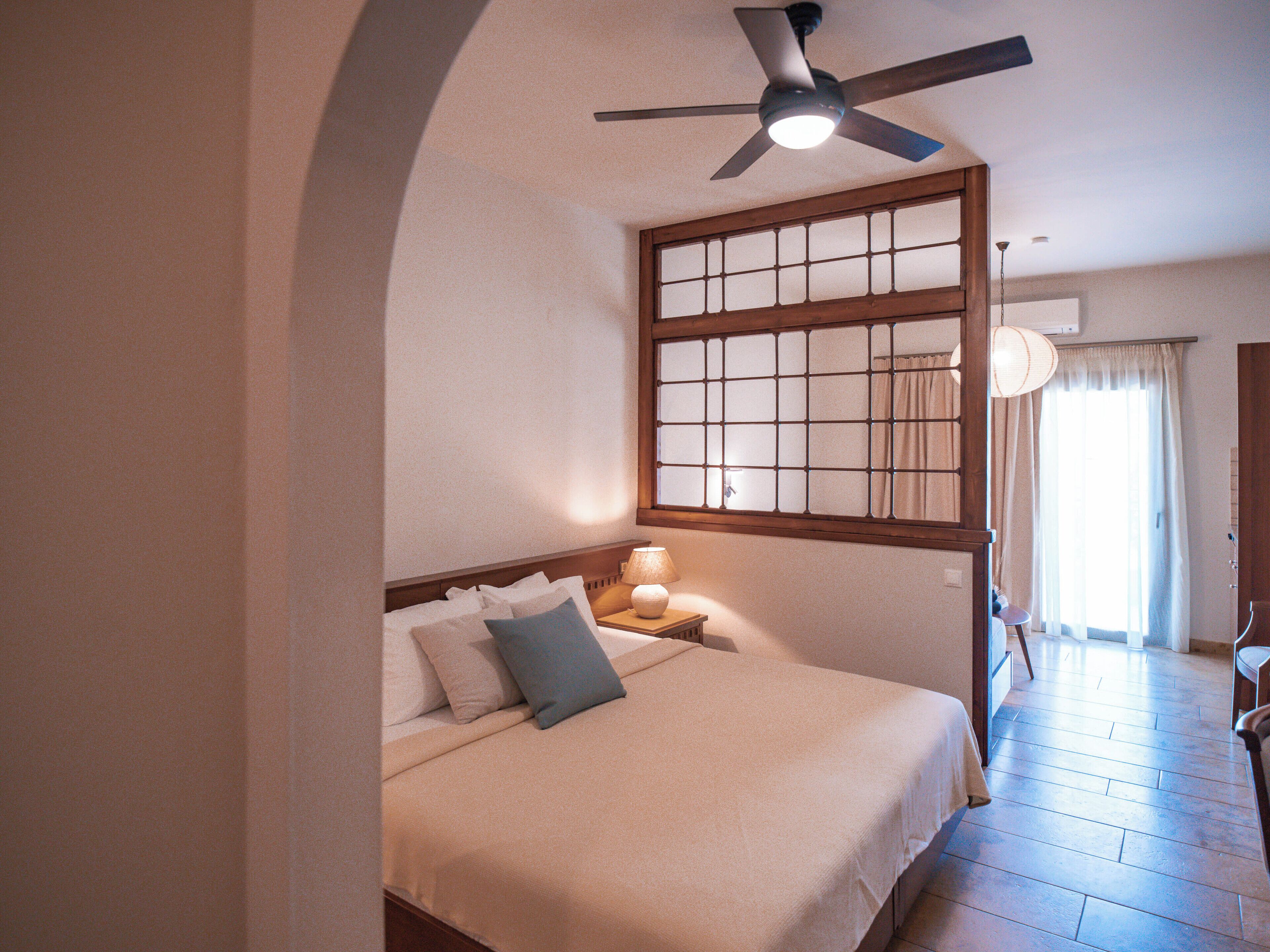 Una habitación de hotel con cama, ventilador de techo, ventana con vistas y mesita de noche con lámpara.