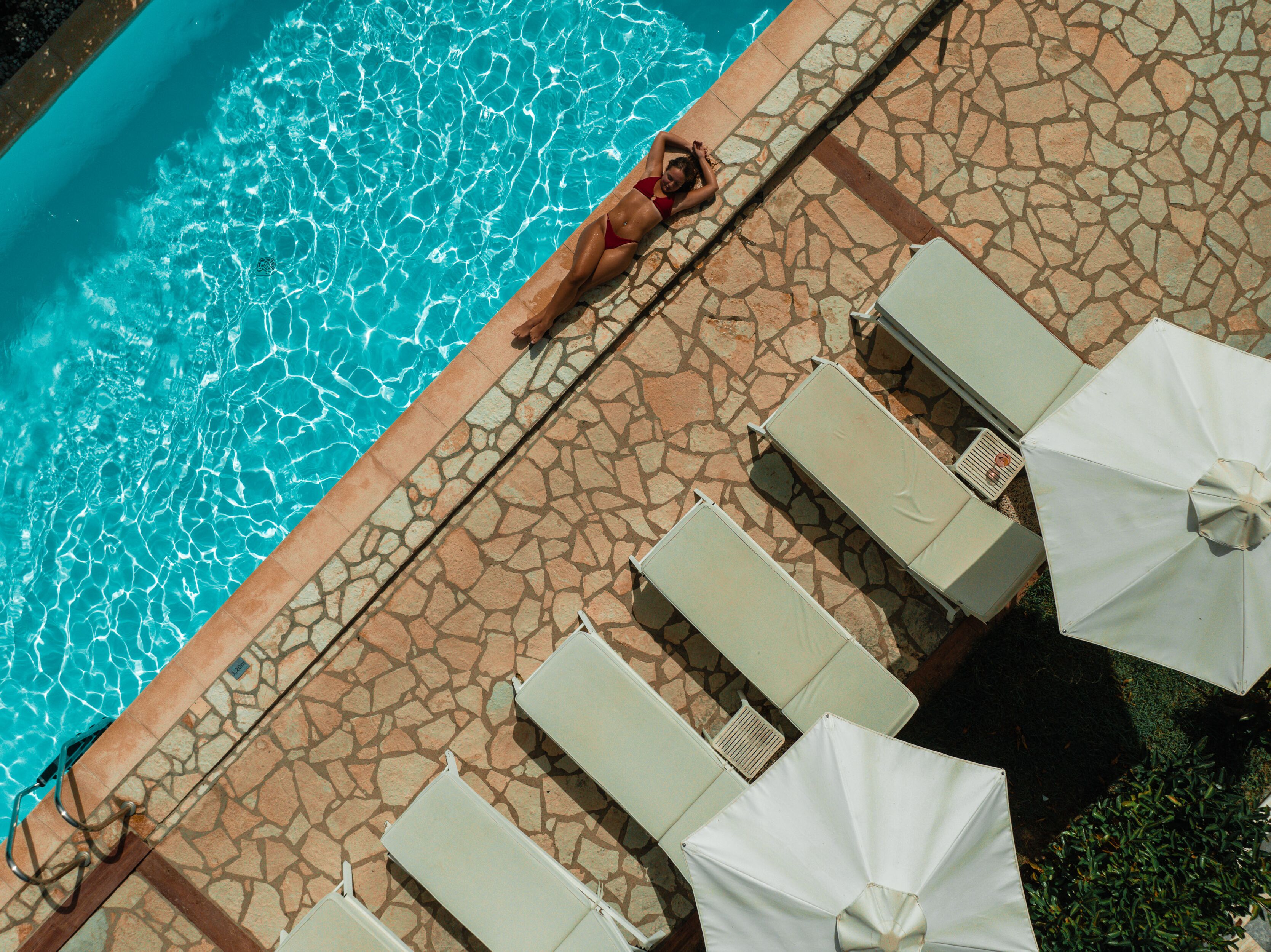 A poolside area with lounge chairs and umbrellas, a tiled walkway, and a person lying on the edge of the pool.