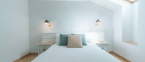 A bedroom with a sloped ceiling, a skylight, a bed with a white mattress and multiple pillows, two bedside tables with lamps, and a small shelf.