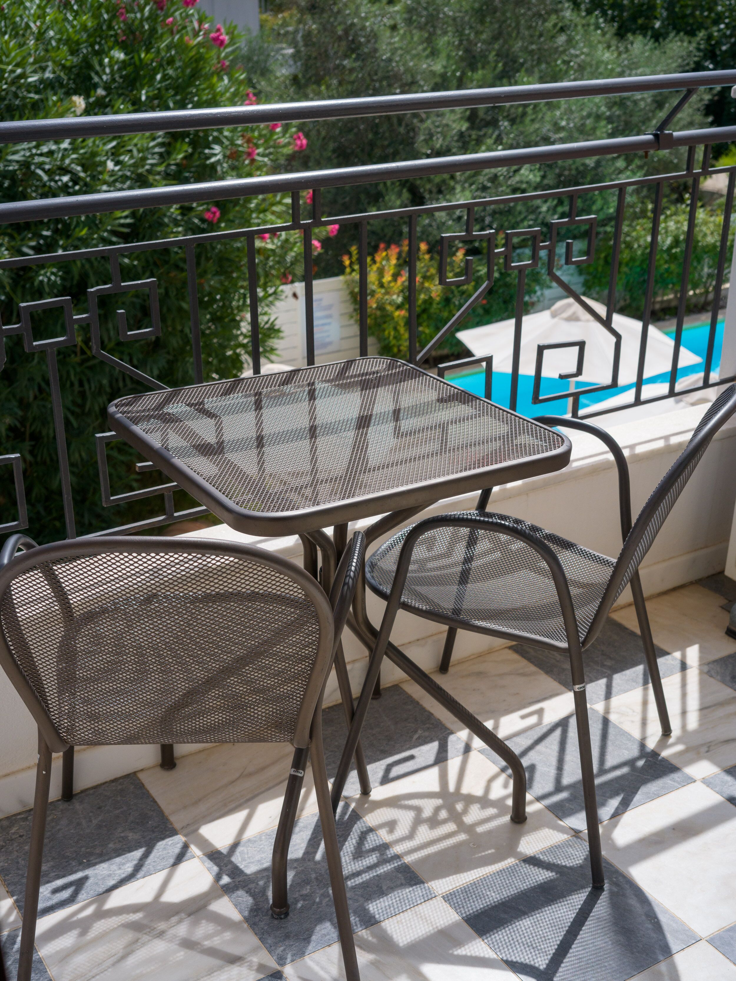 A balcony with a metal table and chairs, a pool in the background, and a tiled floor.