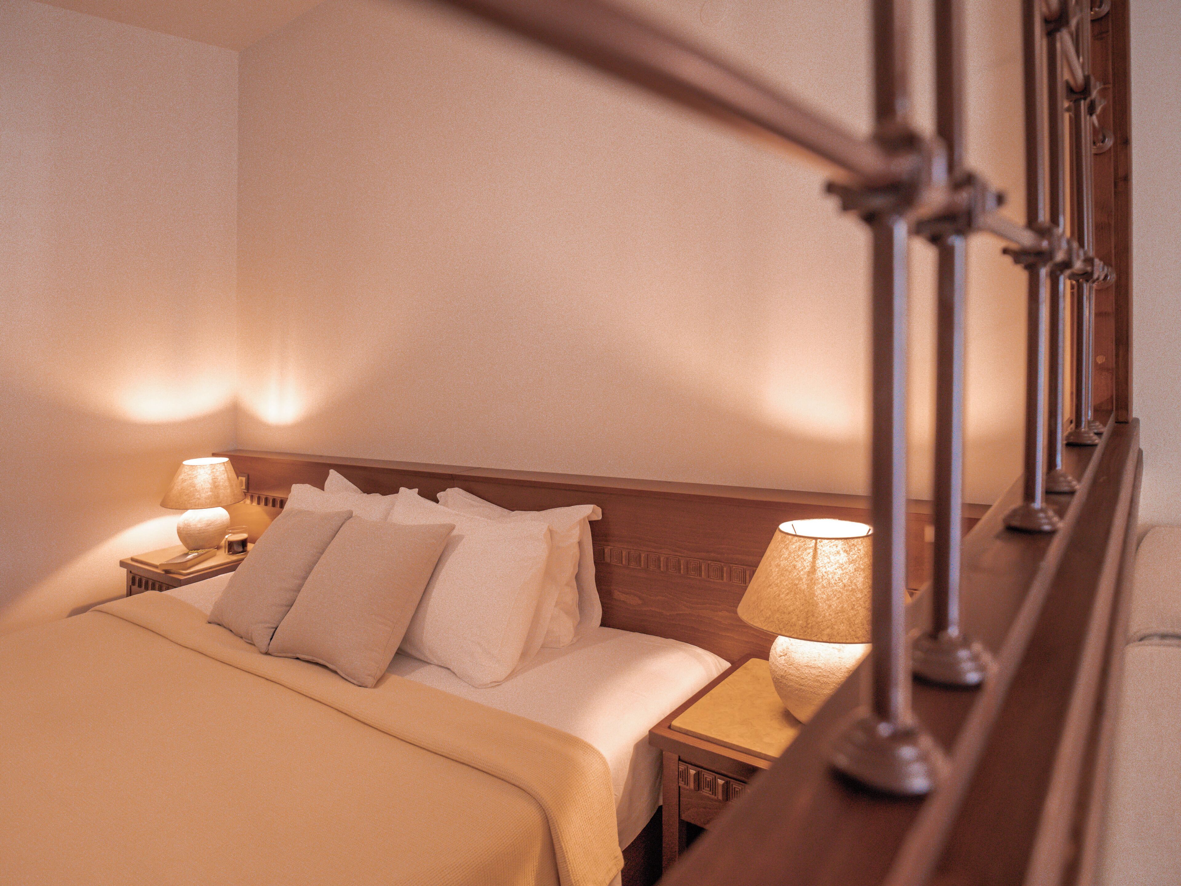 Una cama bien hecha con tablero de madera, dos mesitas de noche con lámparas y una mesita auxiliar de madera.